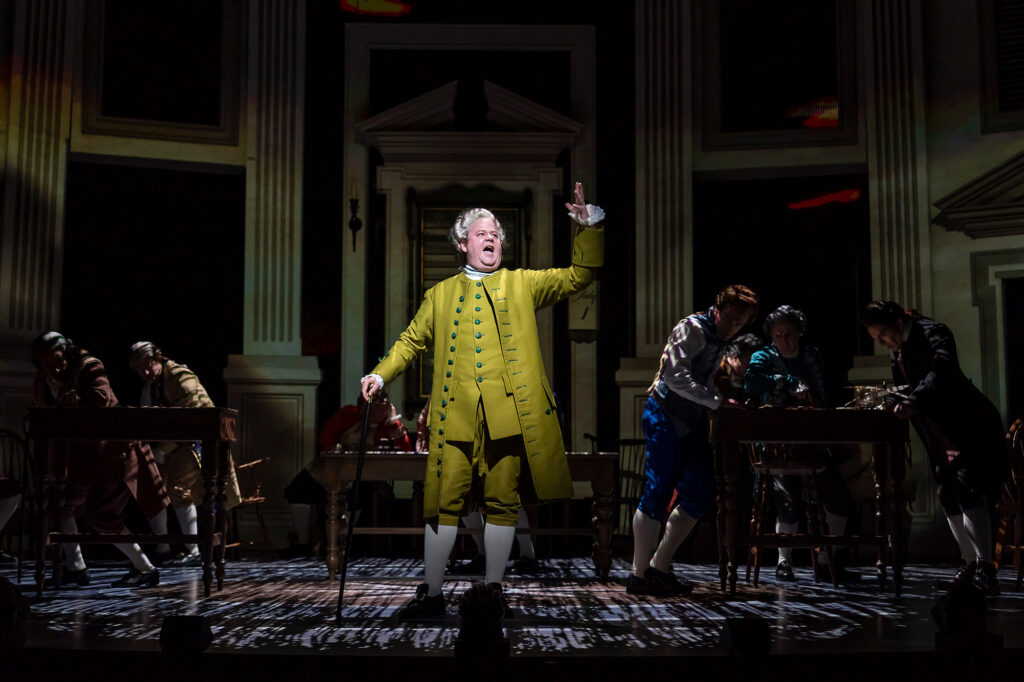 A man in an 18th‑century yellow coat stands center stage, holding a cane and gesturing upward while illuminated by a spotlight. Other actors in period clothing work at desks behind him on a dimly lit set styled like a colonial assembly hall.