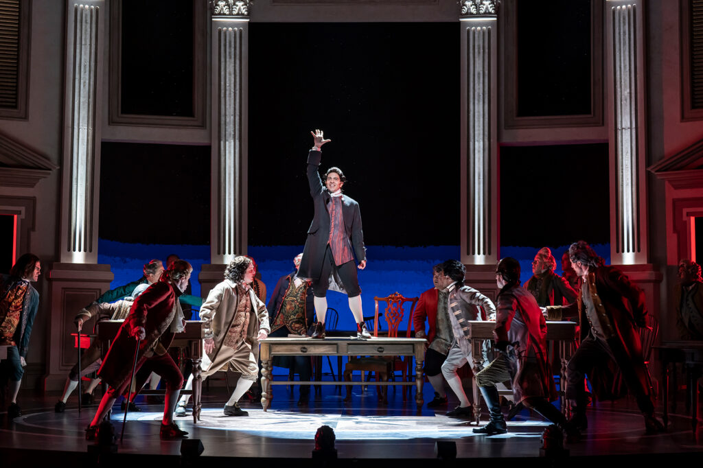 A man in an 18th‑century coat stands triumphantly on top of a table with one arm raised, surrounded by other actors in period costumes who look up toward him. The dramatic lighting and grand colonial‑style set heighten the intensity of the scene.