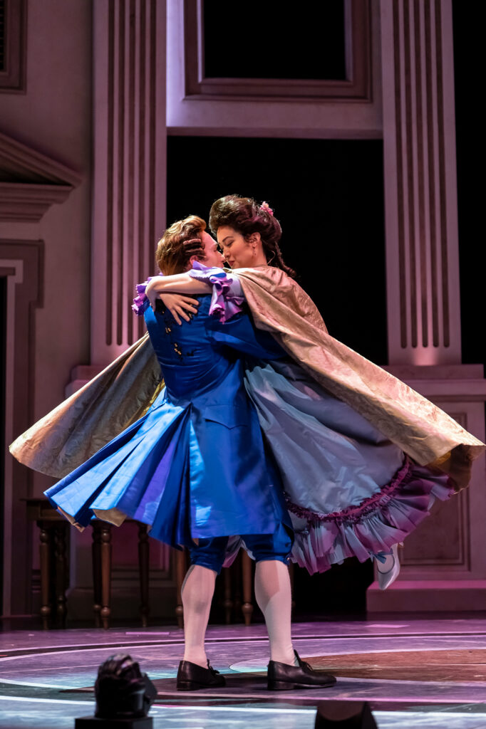 A couple in 18th‑century costumes embrace in a joyful spinning lift onstage. A man in a bright blue coat holds up a woman in a flowing lavender dress and cape, their faces close as they smile in mid‑movement.