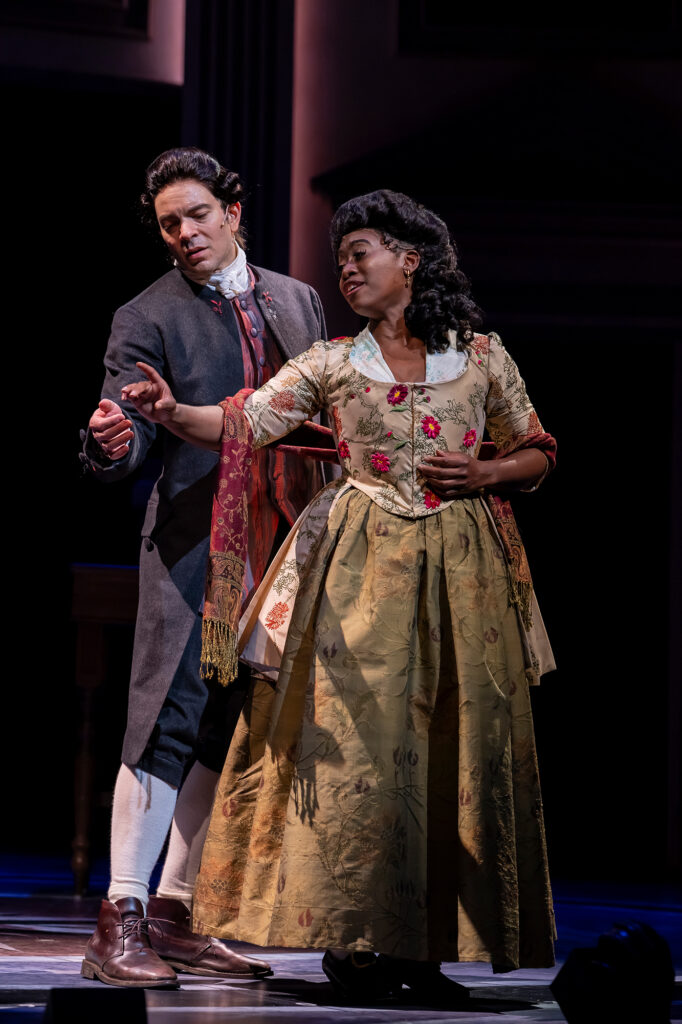 Two performers in elaborate 18th‑century costumes share a gentle, intimate moment onstage. The woman in a floral brocade gown guides the hand of the man beside her, who wears a dark coat and cravat. Both appear mid‑dance in soft theatrical lighting.