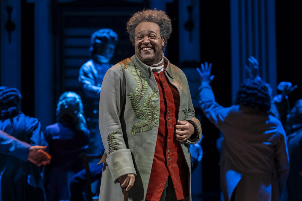 A man in an 18th‑century coat decorated with embroidered green leaves stands smiling at center stage. Warm lighting highlights his expression while other men in darker, blue‑lit costumes gesture in the background.
