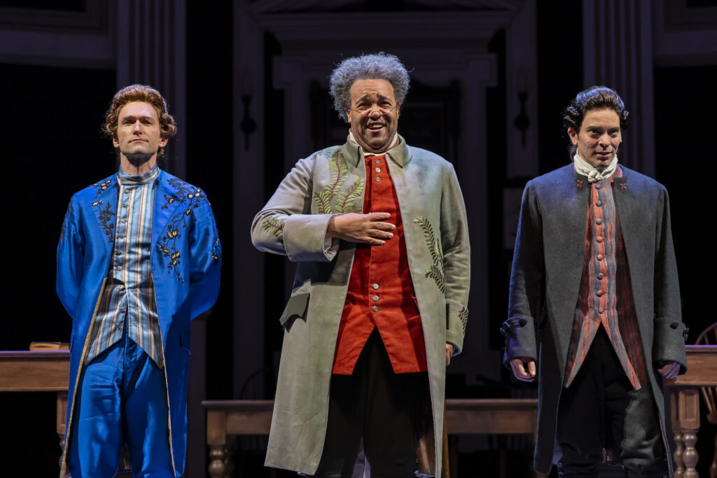 Three men in 18th‑century costumes stand side by side onstage. The man in the middle smiles warmly with a hand on their chest, wearing a light coat with embroidered green leaves. The men on either side wear a bright blue outfit and a dark gray coat, all lit by dramatic stage lighting.