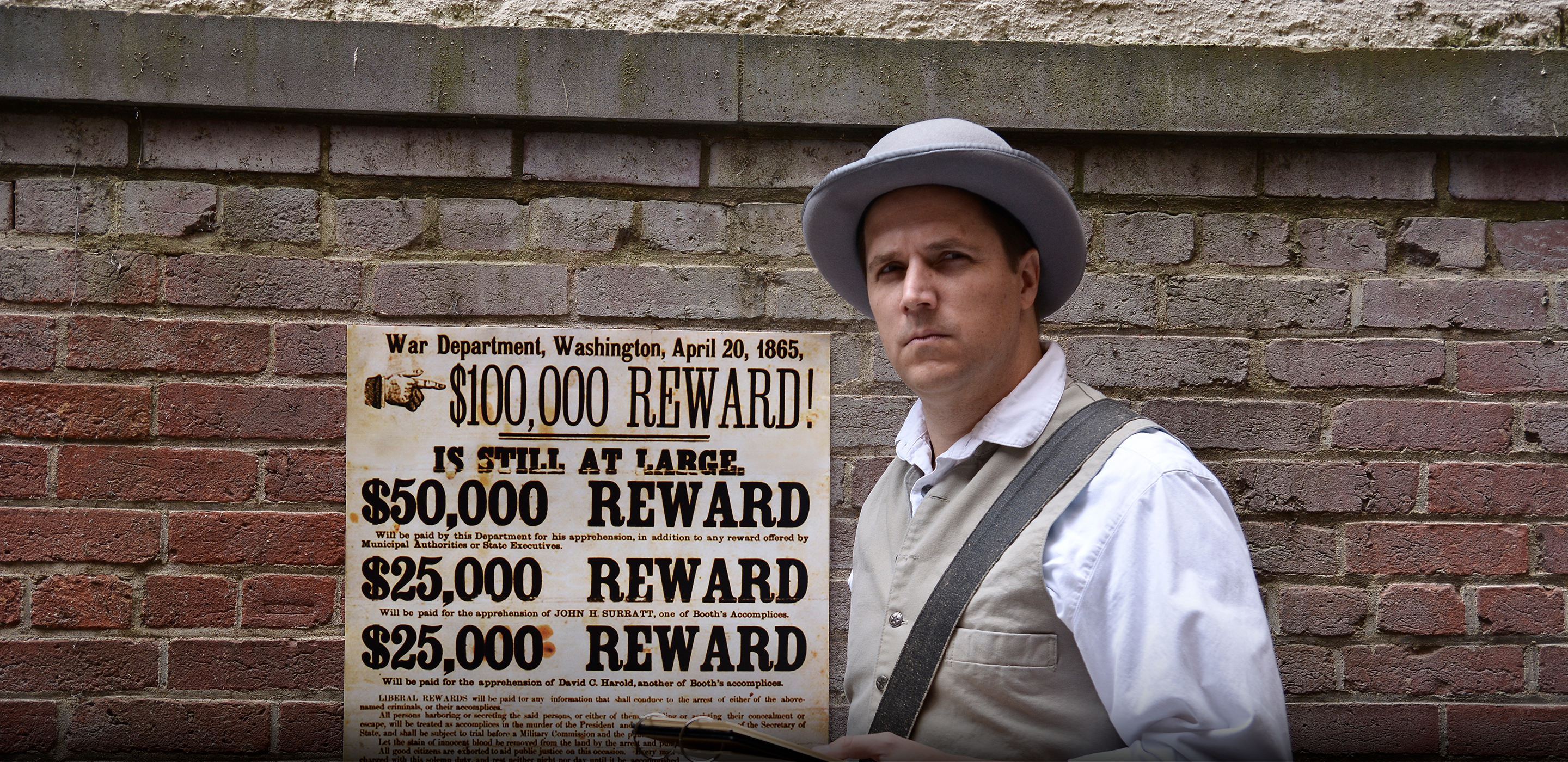 An actor stands dressed as an 1860s detective, holding a notepad. Behind him on a brick wall is posted a reward poster for the capture of Lincoln's assassin.
