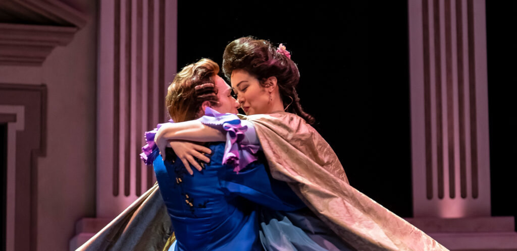A couple in 18th‑century costumes embrace in a joyful spinning lift onstage. A man in a bright blue coat holds up a woman in a flowing lavender dress and cape, their faces close as they smile in mid‑movement.