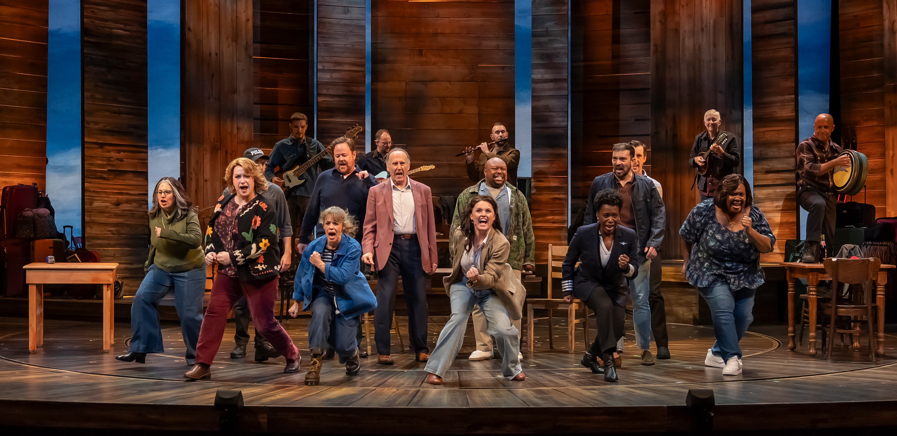 A group of performers and musicians standing across a theater stage, with several musicians playing instruments including an accordion, a fiddle, a bodhrán drum, and guitars. The performers are positioned across the circular wooden floor, stepping and dancing forward with energetic expressions. Tall wooden panels form the backdrop against a bright blue lit sky, illuminated by hanging pendant lights above.