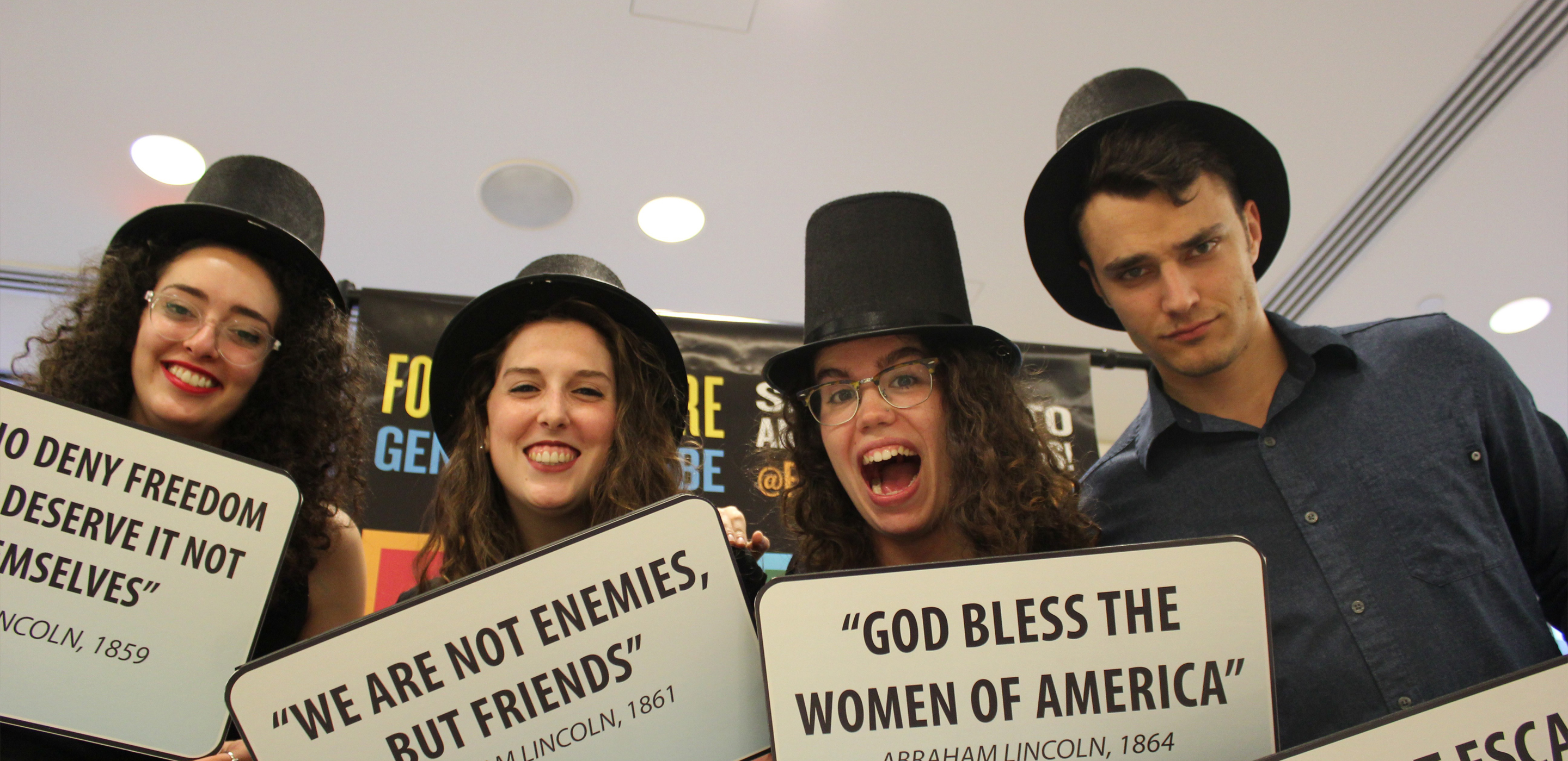 Four young people wearing Lincoln-style top hats hold up placards and smile for the camera.