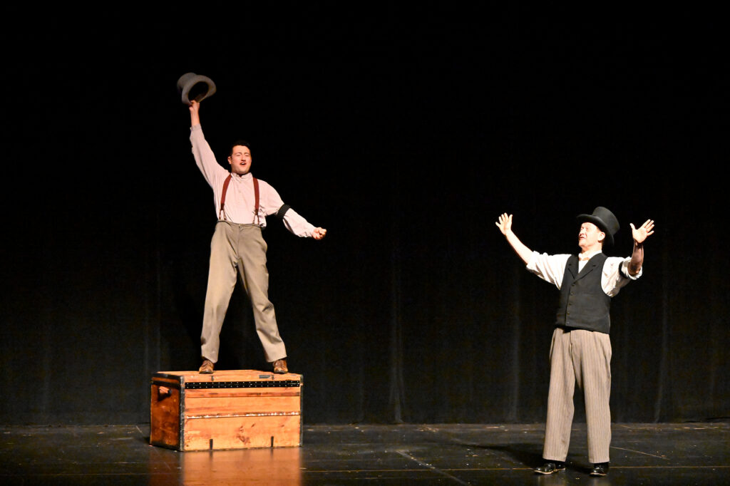 A man in 19th century clothing holding a hat in the air stands on a large chest, while another similarly dressed man holds up his arms.