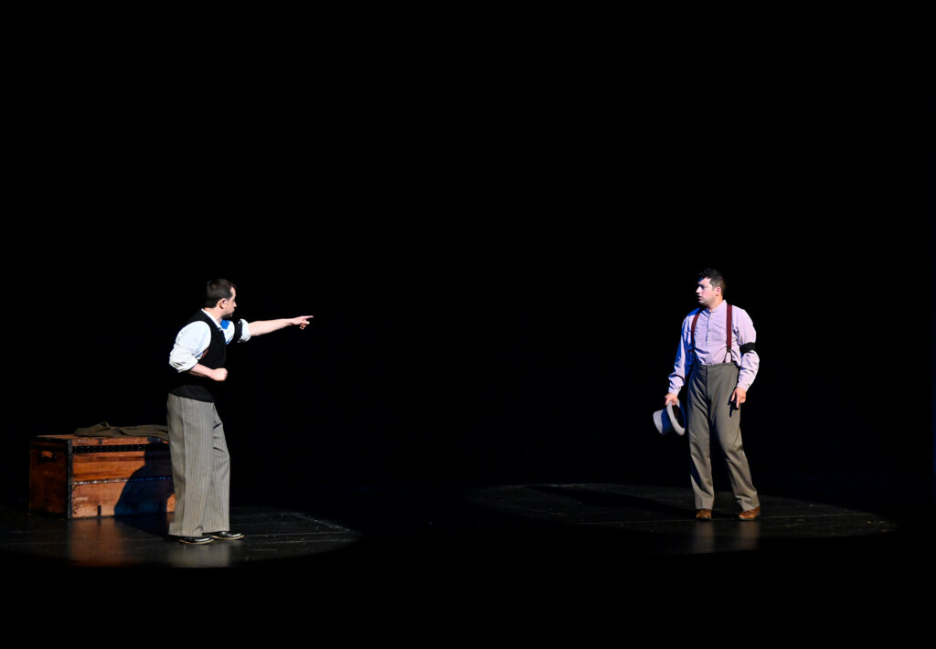 Two men dressed in 19th century clothing stand far apart on a stage. The one on the left is pointing at the one on the right.