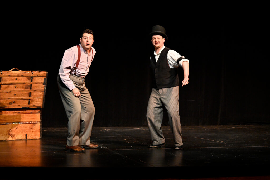Two men dressed in 19th century clothing stand on a stage and look out towards the camera with anxious expressions.
