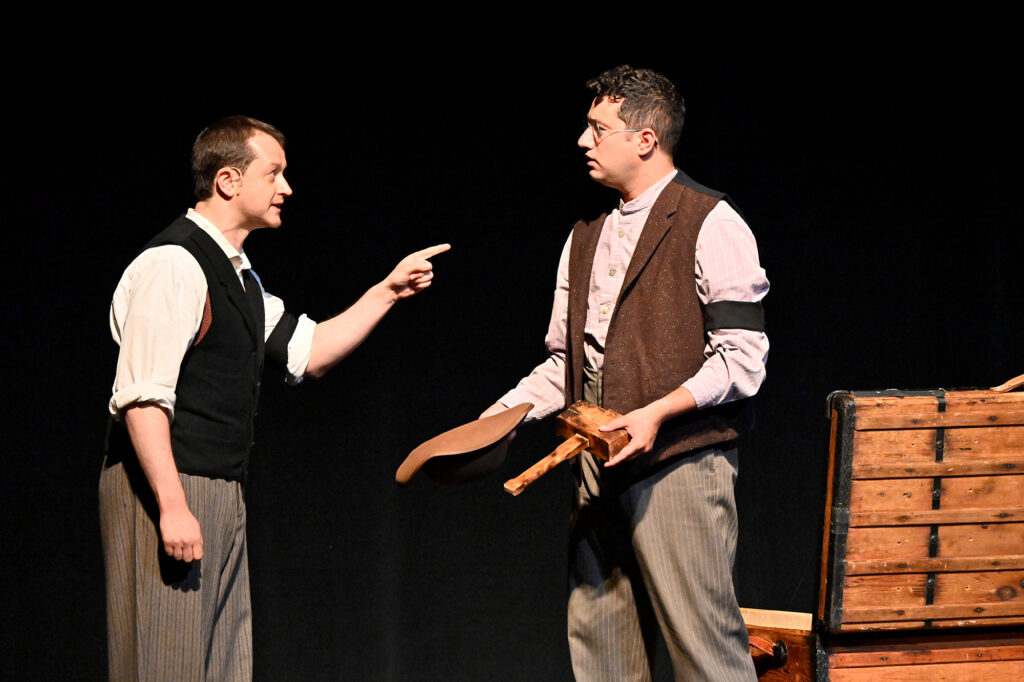 Two men dressed in 19th century clothing stand on a stage. The one on the right is pointing at the one on the left, who holds a wooden mallet and a piece of leather.