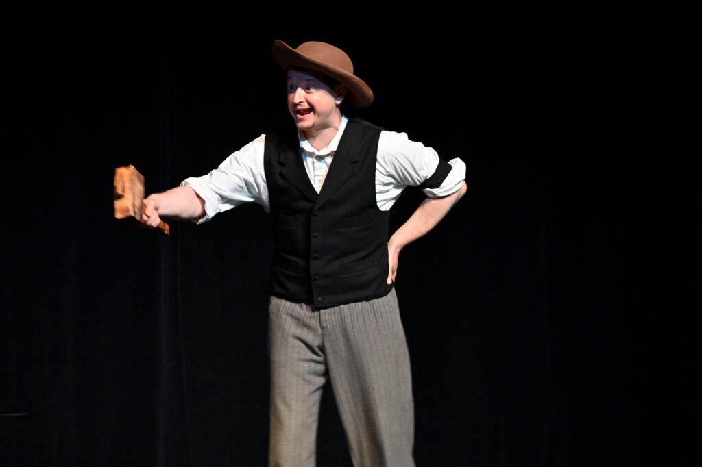 A man in 19th century clothing stands on a stage and gesticulates with a large wooden mallet.