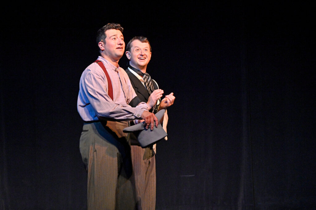 Two men dressed in 19th century clothing stand on a stage and look out towards the audience with happy expressions.