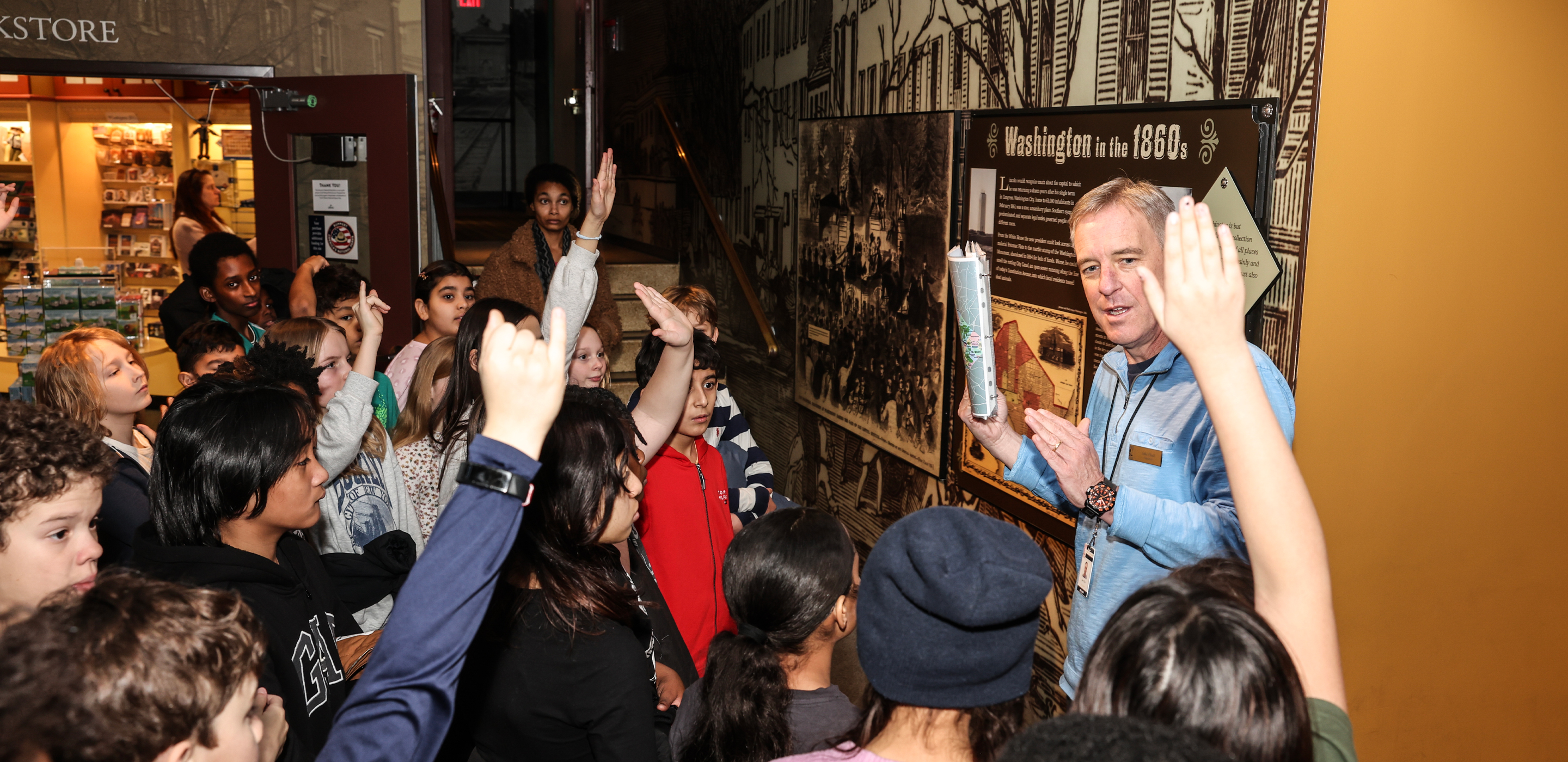 A group of students listen raise their hands while listening to an adult man discuss a museum display.