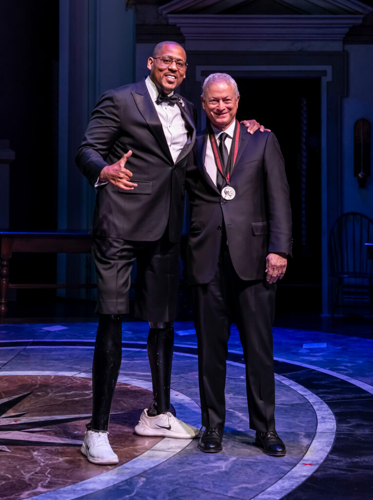 A tall Black man with two prosthetic legs poses with a white man on a stage, both wearing tuxedos.