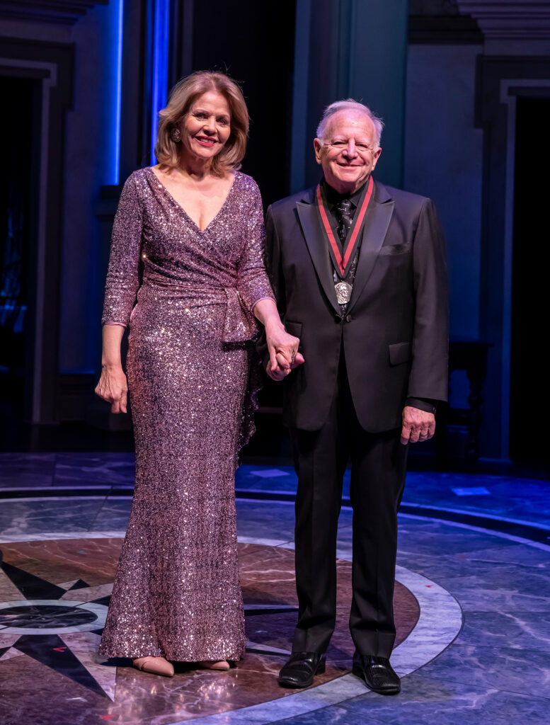 A white woman in a purple dress stands next to an older white man in a suit on a stage.