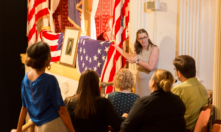 A woman stands in the Ford’s Theatre balcony near the Presidential Box leading a guided tour for seven people who are seated.