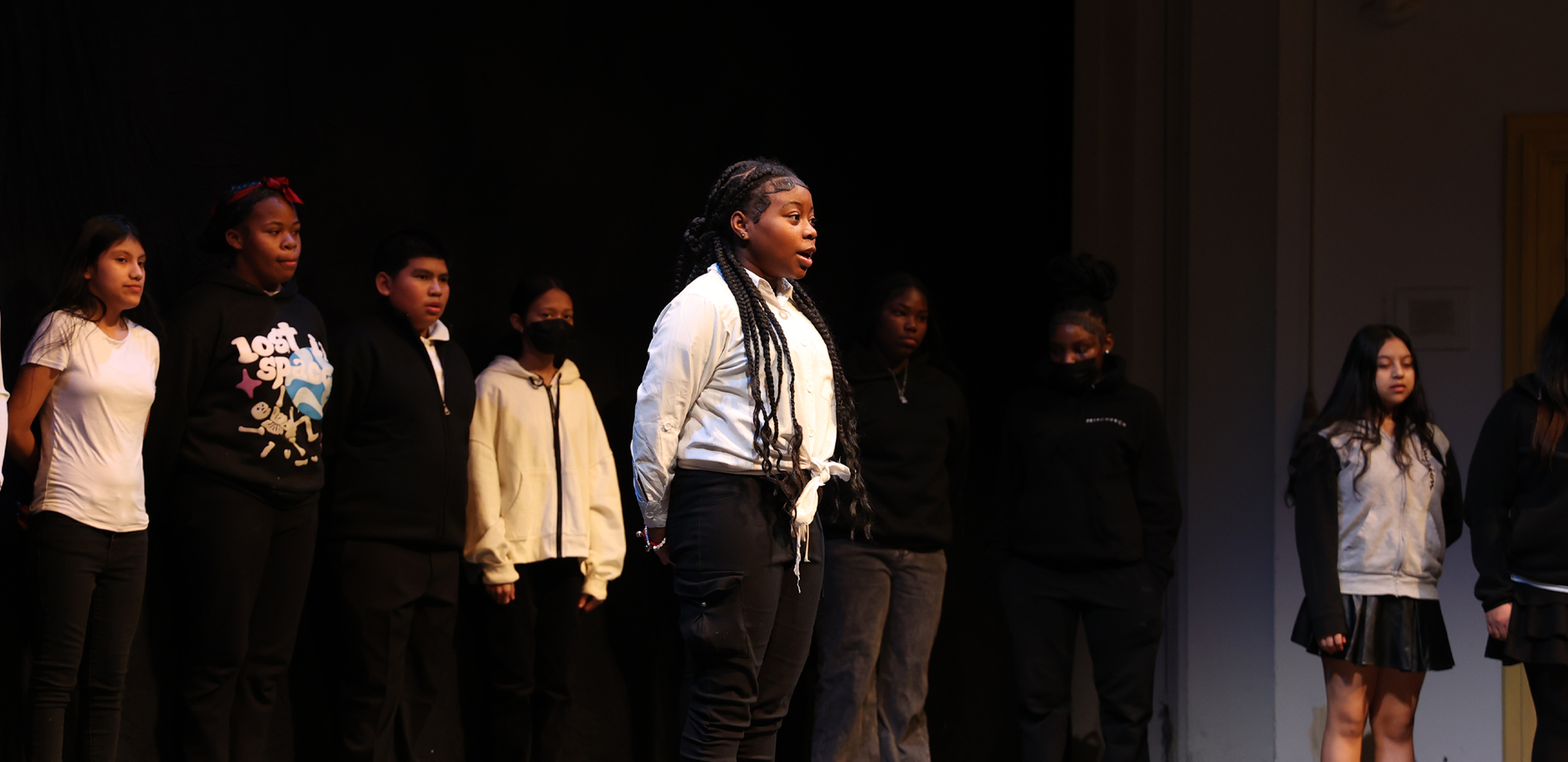 A female student stands center stage performing spoken word as her peers watch on in support in the background.