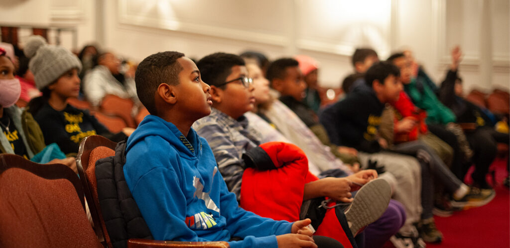 A group of young students sit in the audience of a theatre.