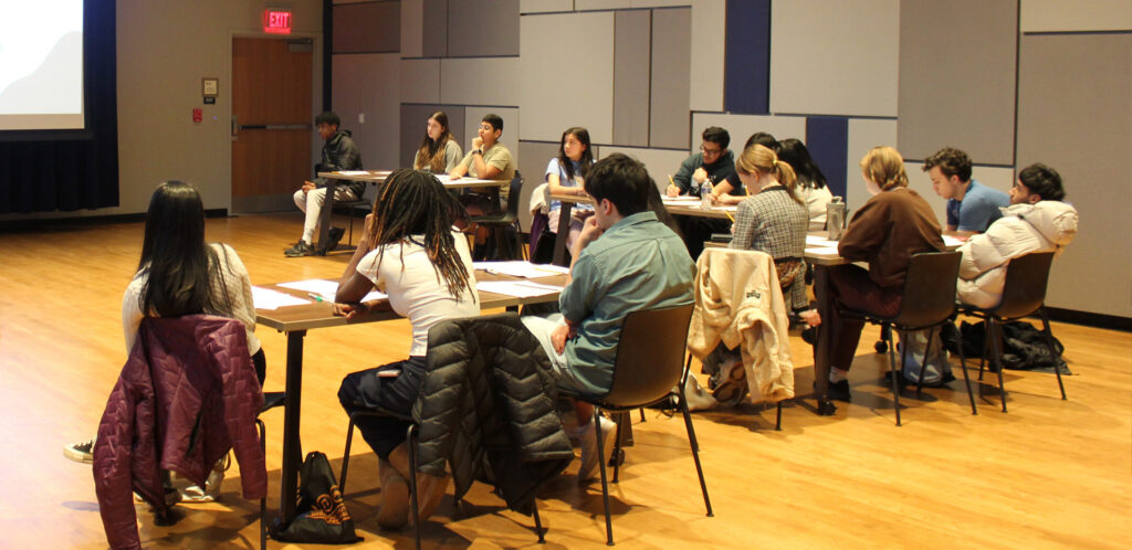 A group of students sit at tables in a large room and watch a presentation on a screen.