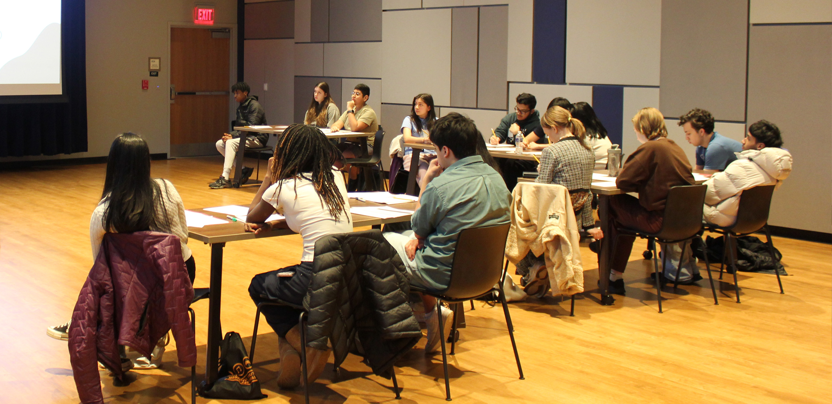A group of students sit at tables in a large room and watch a presentation on a screen.