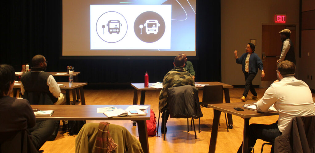 A group of men and women sit at tables in a large studio and watch a presentation projected onto a screen.