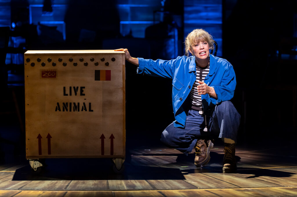 A woman crouches on a wooden stage, resting one hand on a large wooden shipping crate stamped with the words "LIVE ANIMAL" and a Belgian flag. She wears a bright blue jacket, a striped shirt, and wide-leg jeans, looking up with an earnest look under a cool blue spotlight.