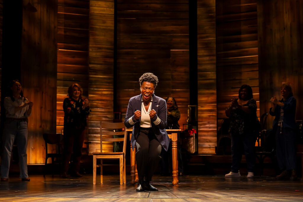 A woman in a pilot’s jacket and trousers stands at center stage, crouched forward with clenched fists and a triumphant expression as she sings. Behind her, other performers stand in the background, smiling and applauding under warm, glowing lighting against a backdrop of tall wooden panels.