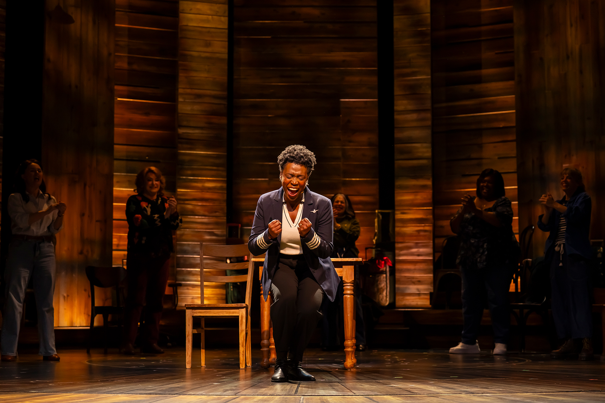 A woman in a pilot’s jacket and trousers stands at center stage, crouched forward with clenched fists and a triumphant expression as she sings. Behind her, other performers stand in the background, smiling and applauding under warm, glowing lighting against a backdrop of tall wooden panels.