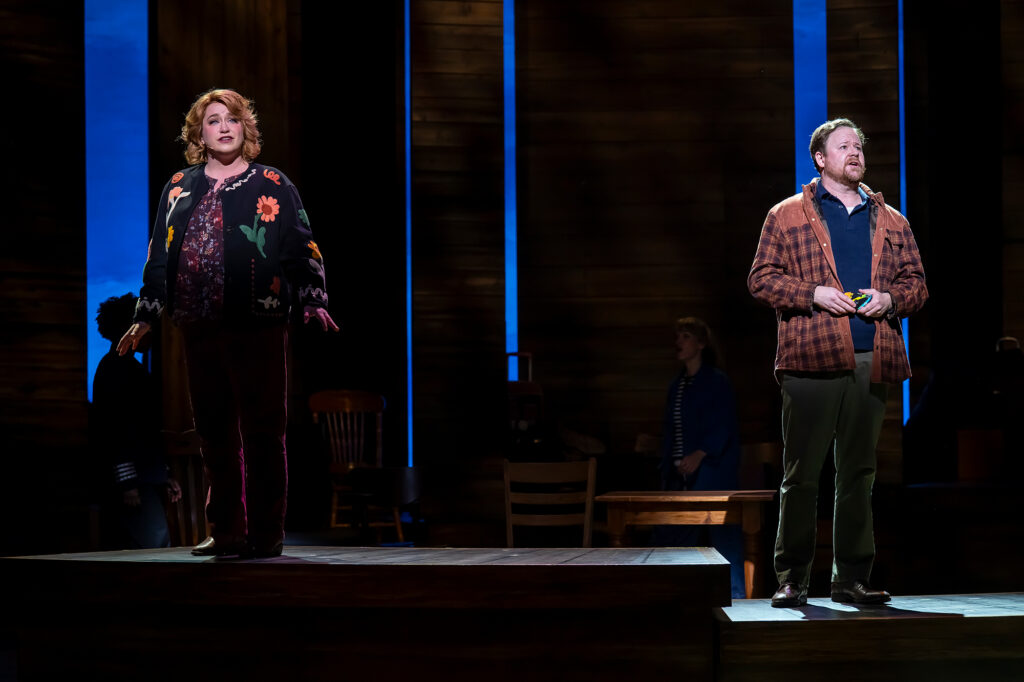 A woman and a man standing apart on a wooden stage, both looking out toward the audience while singing. On the left, the woman wears a floral embroidered cardigan sweater and holds her hands slightly outward with an emotional expression. On the right, the man stands in a plaid jacket and polo shirt, holding a small camera in his hands, set against tall wooden backdrop panels and soft blue lighting.