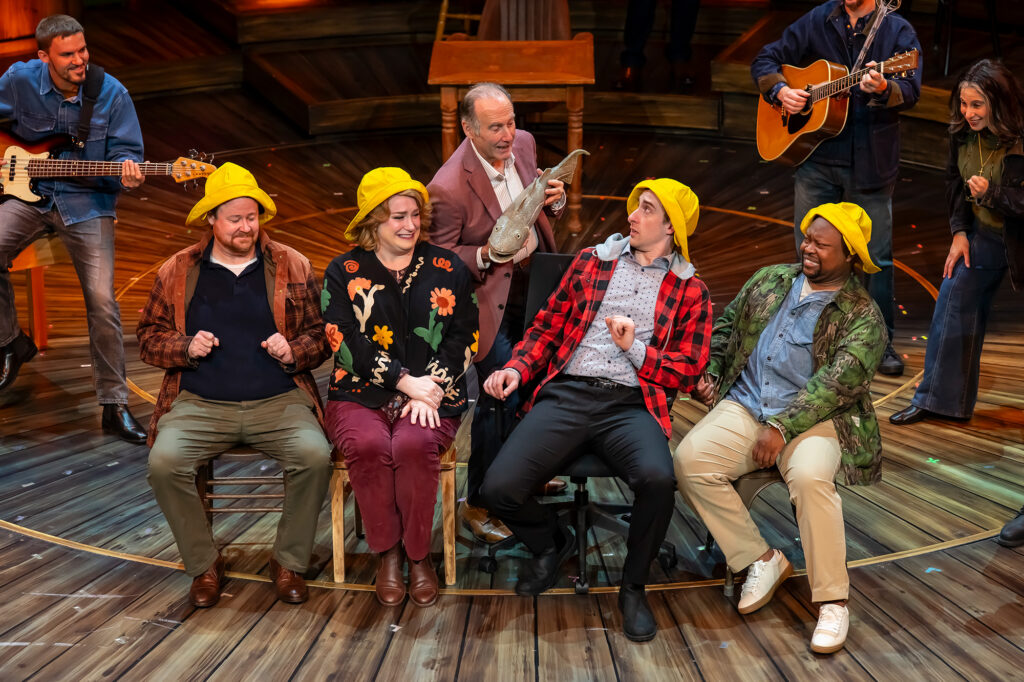 Four performers seated in a row on chairs across a circular wooden stage, each wearing a bright yellow fisherman's hat. Behind them, a man in a blazer holds up a large fish prop toward the seated cast, while musicians on either side play a bass guitar and an acoustic guitar under warm stage lighting.
