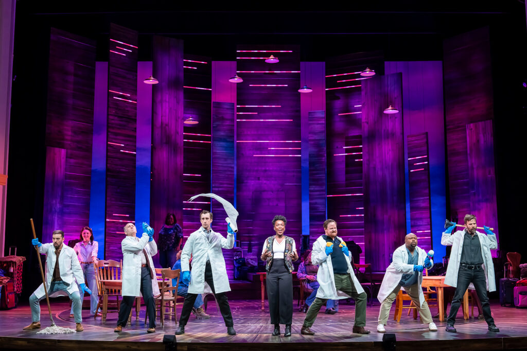 Performers on stage bathed in deep purple and magenta light. Several cast members wear white lab coats and blue gloves, striking exaggerated poses while holding cleaning tools such as mops, sponges and a toilet plunger. At center stage, a man and a woman in everyday clothes stand, surrounded by the animated group under overhead lights and tall wooden set panels.