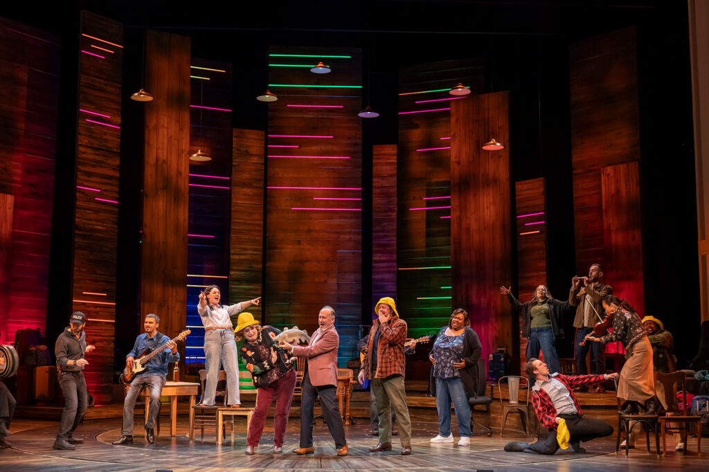 Performers and musicians spread across a theater stage. Near center stage, a man in a blazer holds up a large fish next to a woman in a bright yellow hat, while other cast members cheer, dance, and play instruments, including a bass guitar, a whistle, and a fiddle. Tall wooden backdrops with horizontal neon light strips create a vibrant setting under hanging pendant lights.
