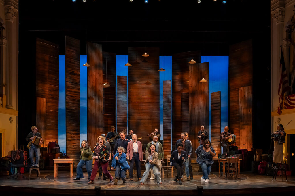 A group of performers and musicians standing across a theater stage, with several musicians playing instruments including an accordion, a fiddle, a bodhrán drum, and guitars. The performers are positioned across the circular wooden floor, stepping and dancing forward with energetic expressions. Tall wooden panels form the backdrop against a bright blue lit sky, illuminated by hanging pendant lights above.