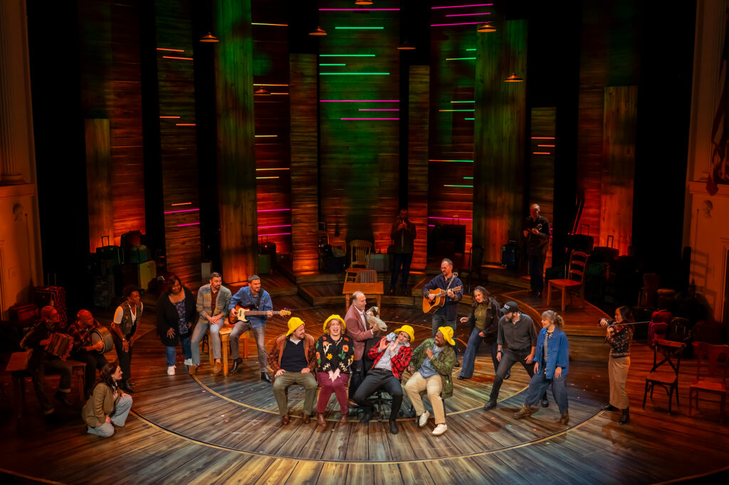 A large group of performers gathered on a theater stage, with several seated in the center wearing colorful clothing and yellow hats while others stand or sit around them. Musicians play guitar and other instruments among the group. The stage features a circular wooden floor and tall, staggered wooden panels in the background, illuminated with colorful horizontal strips of green, pink, orange and yellow light. Chairs, tables and luggage are arranged throughout the set.