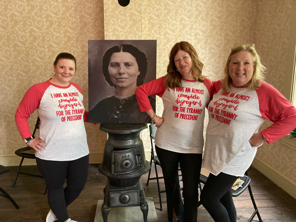 Three women in tee shirts that read "I have an almost complete disregard for the tyranny of precedent" stand next to a black and white photograph of a woman in 19th century clothing.