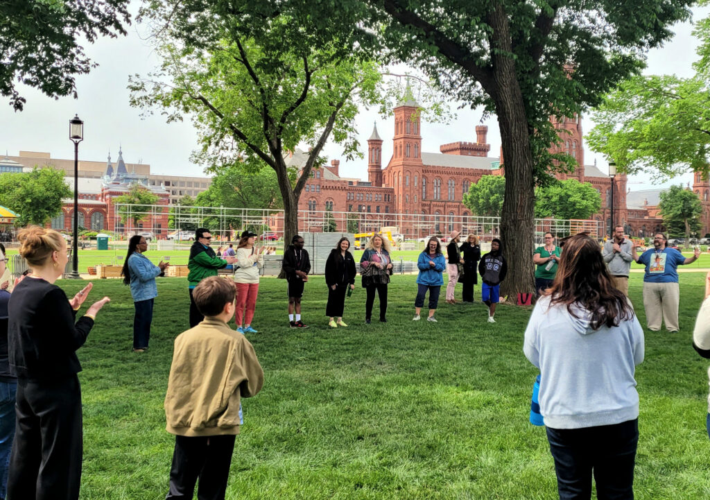 A large group of people stand in a circle on the lawn in front of the Smithsonian Castle.