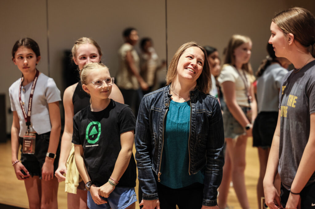 A woman stands in a rehearsal room and interacts with a group of teenage girls.