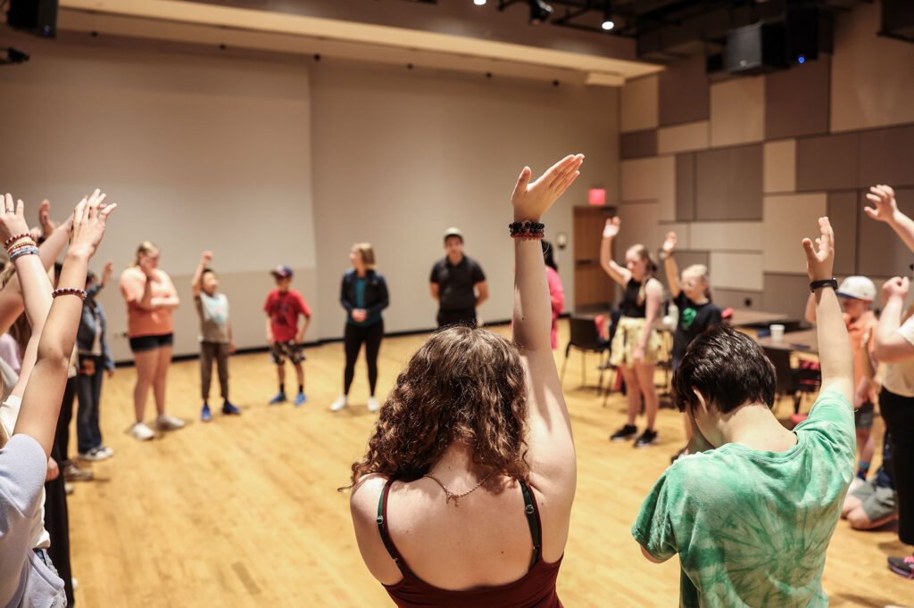 A group of people stand in a circle in an auditorium, many with their hands raised.