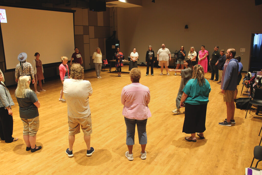A group of people stand in a circle in a rehearsal room. An instructor stands in the middle of the circle and points.