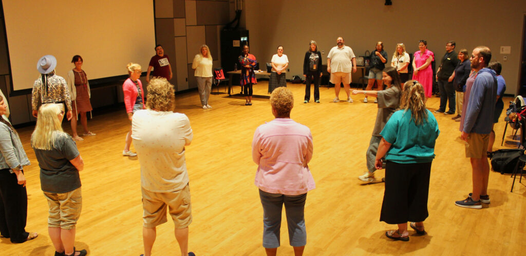 A group of people stand in a circle in a rehearsal room. An instructor stands in the middle of the circle and points.