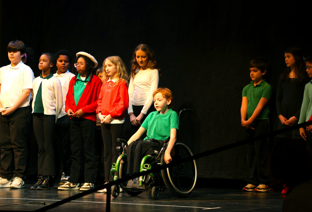 A boy in a wheelchair wearing a green shirt speak while in front of a row of students on a stage.