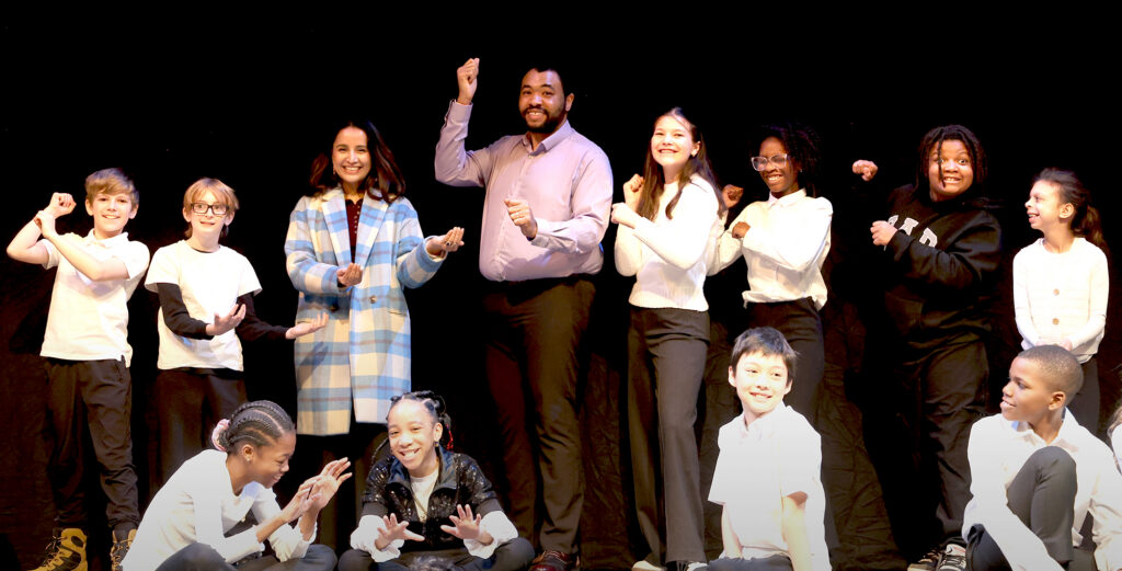 Two adults and a dozen children make cheerful poses while speaking on a stage.
