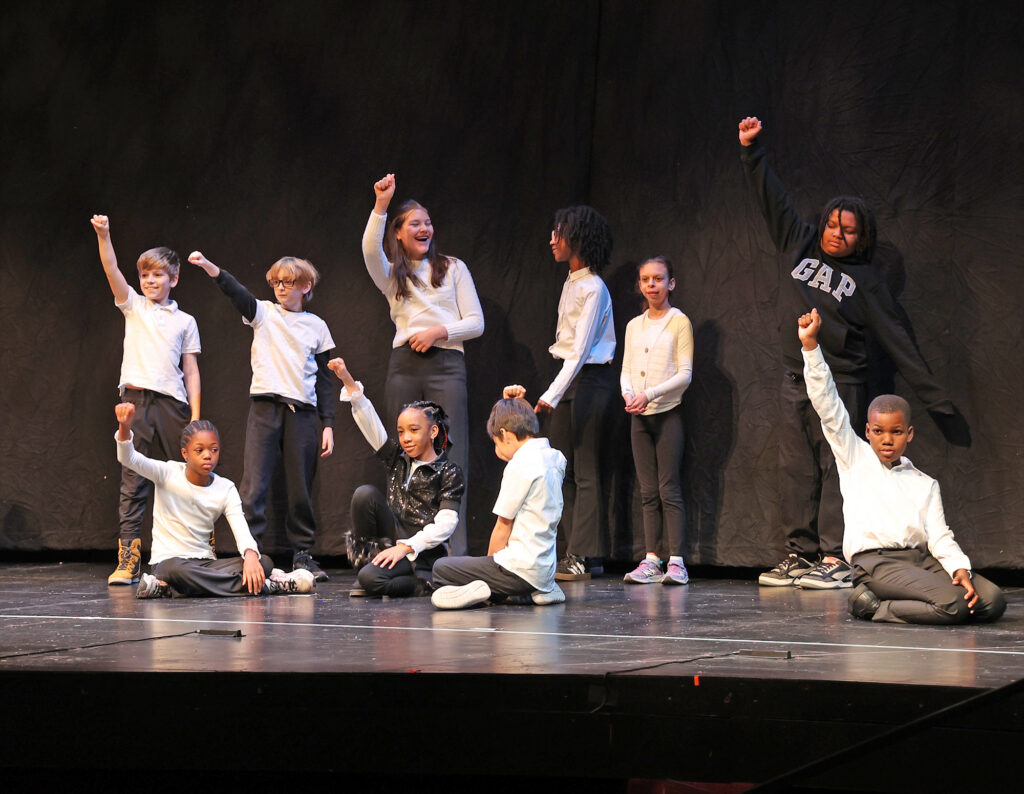 Two adults and a dozen children make cheerful poses while speaking on a stage.