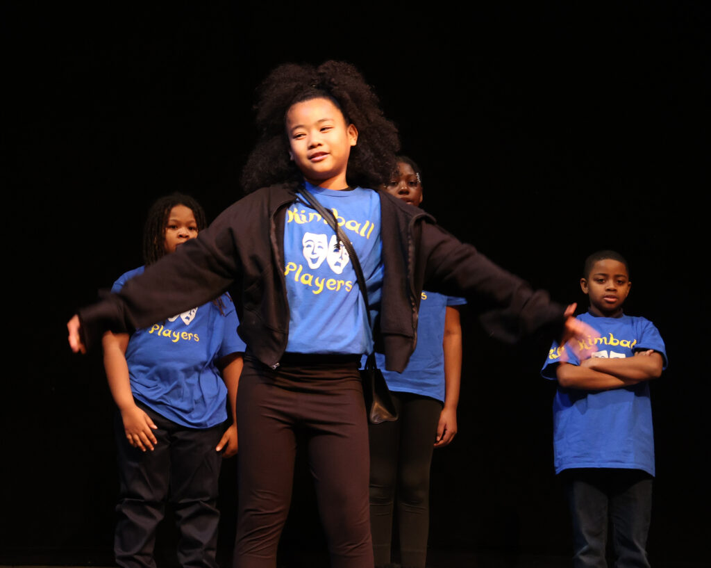 A young girl in a black jacket and blue shirt spreads her arms and speaks on a stage.