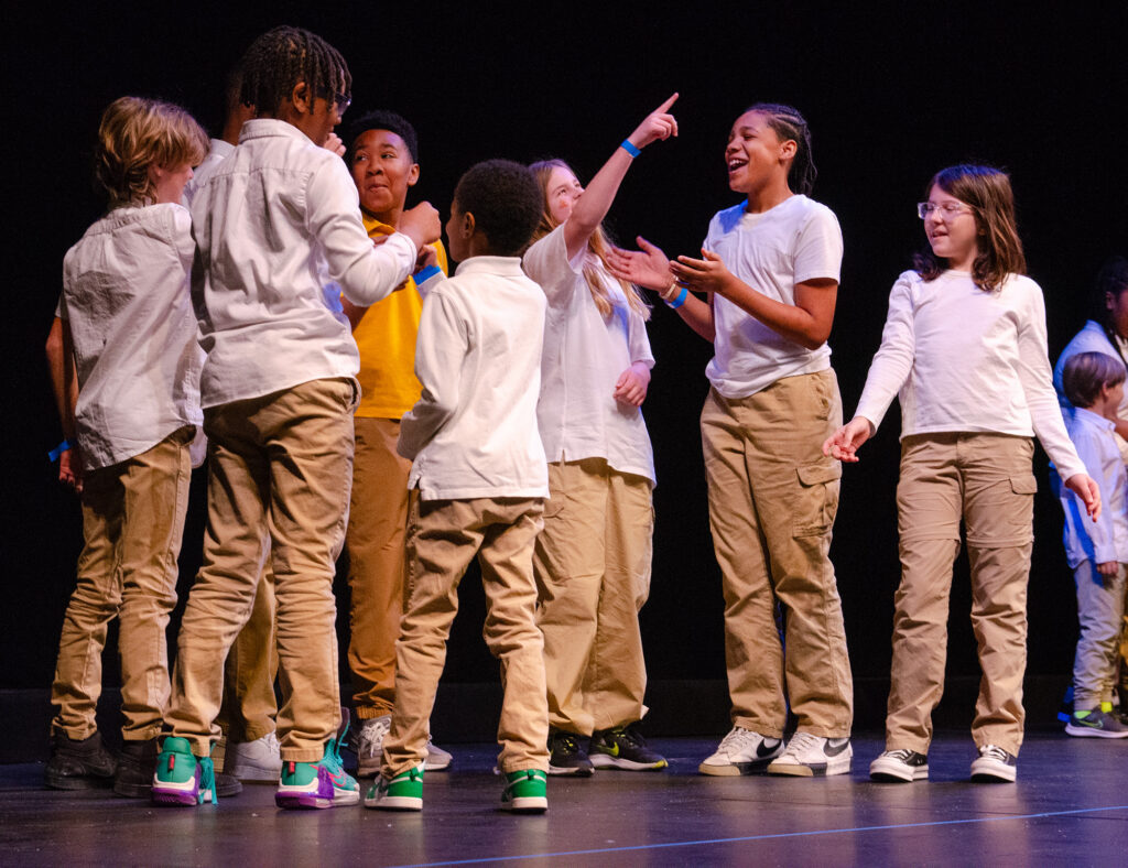 A group of children in white shirts and khaki pants mill about on a stage.
